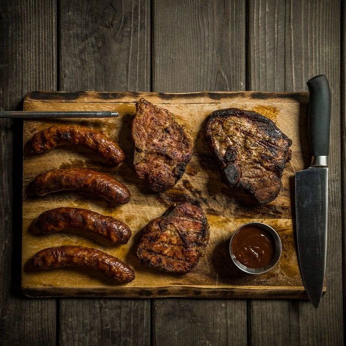 Grilled meats including sausages and steaks arranged on a wooden cutting board, accompanied by a knife and a small bowl of barbecue sauce.