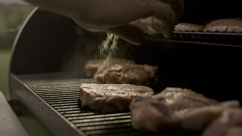 Berkwood+Farms+IA+-+Berkwood+-+4th+-+6-L (1) Close-up of a hand sprinkling seasoning on grilled steaks on a barbecue, with smoke rising and a blurred background of cooking bread, highlighting outdoor grilling techniques.