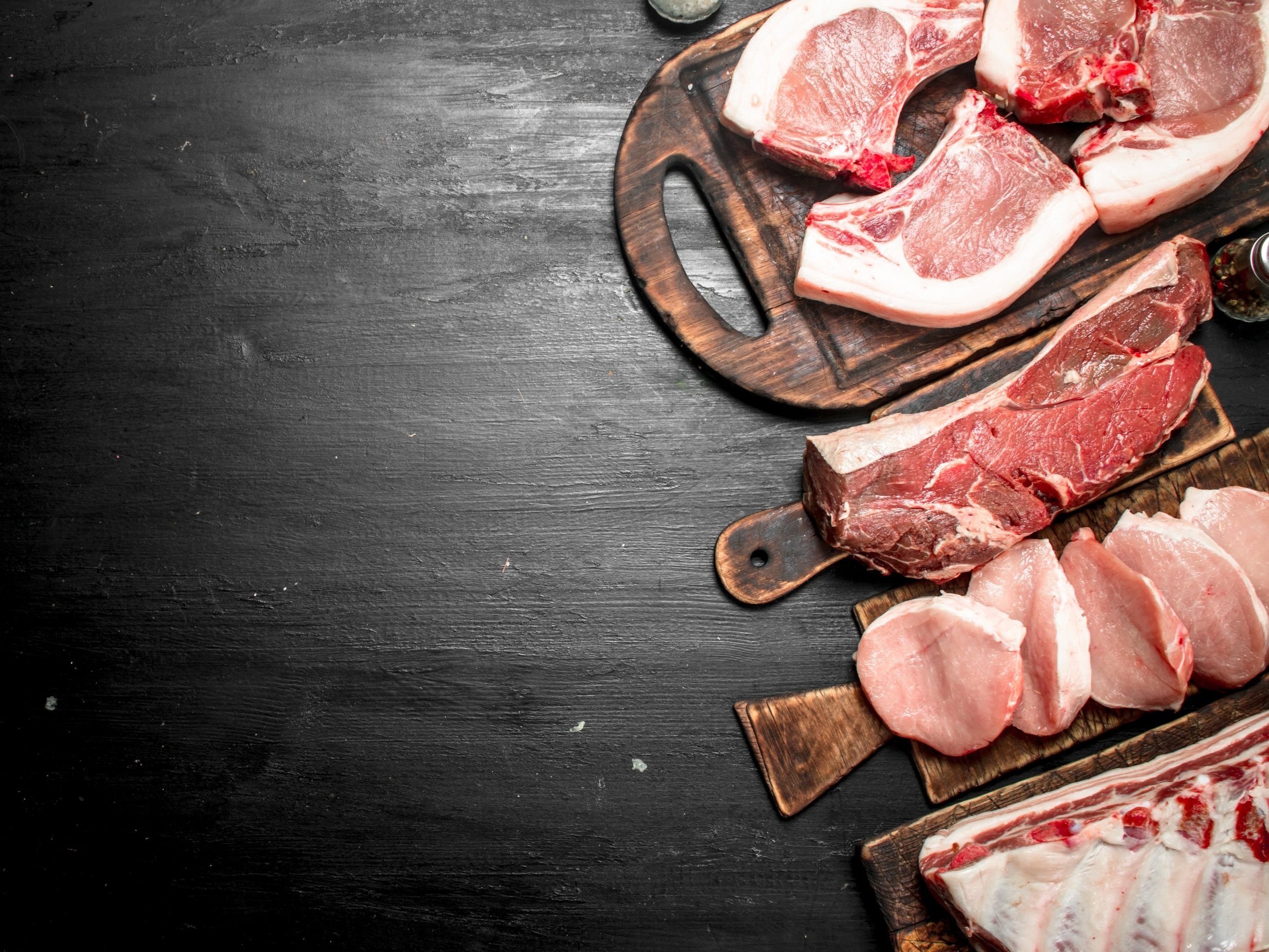 Assorted cuts of fresh meat displayed on wooden cutting boards, including pork chops, beef steaks, and ribs, arranged on a dark background for a culinary presentation.