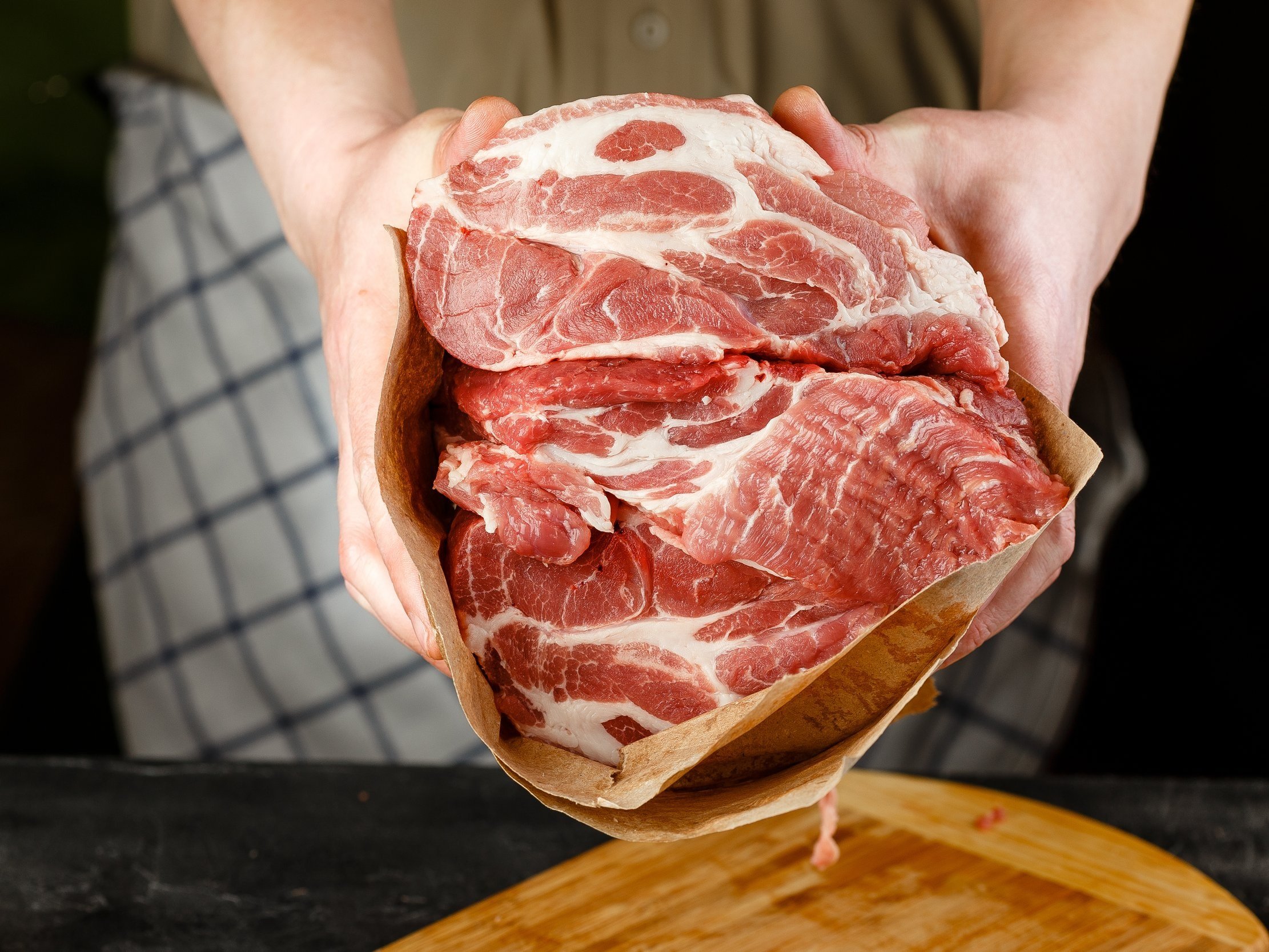 Fresh cuts of pork shoulder held in hands, wrapped in butcher paper, with a wooden cutting board in the background, ideal for cooking and grilling recipes.